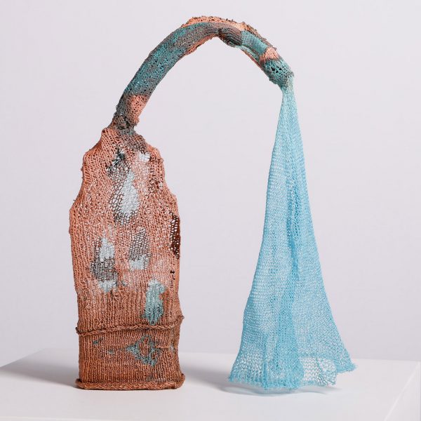 A photograph of a textiles installation in an abstract shape, in tones of blue, red and orange, standing on a white plinth against a white background.