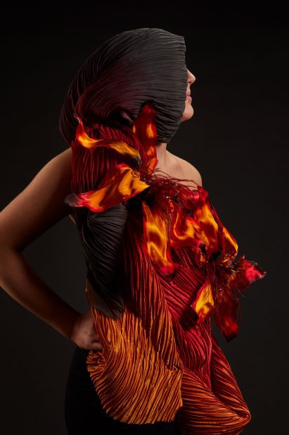 A photograph of a person turned to the side with their hand on their hip, wearing a garment that covers their head and body, in red, orange and black fabrics, against a black background.