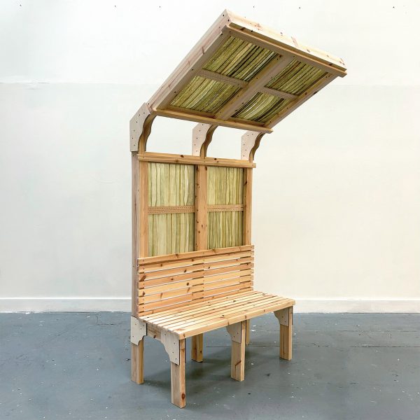 A photograph of a wooden seating structure with an angled shelter over it, made from wood and green seaweed.