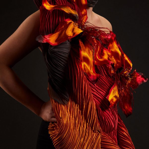 A photograph of a person turned to the side with their hand on their hip, wearing a garment that covers their body, in red, orange and black fabrics, against a black background.