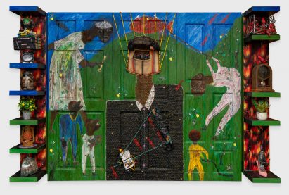 A photograph of a wooden structure that is painted in colours of green, blue, red and yellow, depicting an abstract scene with figures. There are small shelves either side of the strucutre with various objects on them.