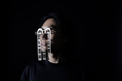 A photograph of a person's head, against a black background, wearing silver metal glasses that replicate architectural details.
