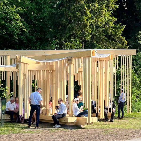 A photograph of an outdoor wooden structure in tones of beige, amongst green trees. There are people underneath it.