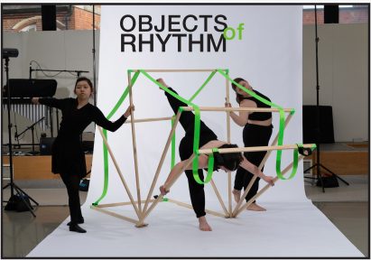 A photograph of three people dressed in black handing a wooden structure with green rope around it. In the centre of the image, above the people, are the words 'objects of rhythm' written in black and green.