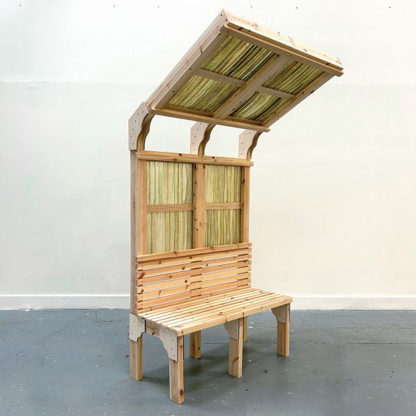 A photograph of a wooden seating structure with an angled shelter over it, made from wood and green seaweed.