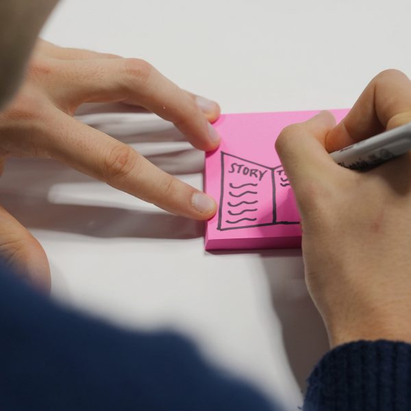 A photograph of a person's hand writing on a pink post it note.