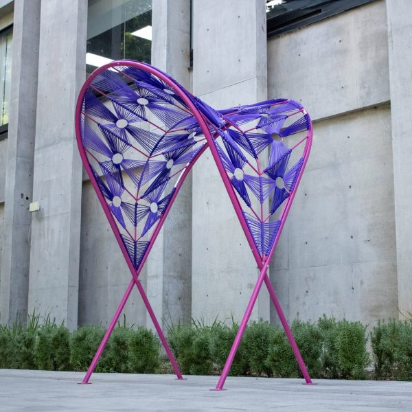 A photograph of an outdoor installation in tones of purple and pink against a grey concrete wall.