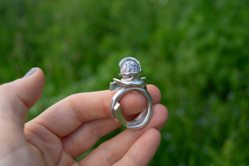 Photo of a person's hand holding a silver ring topped with a small orb above a grassy field
