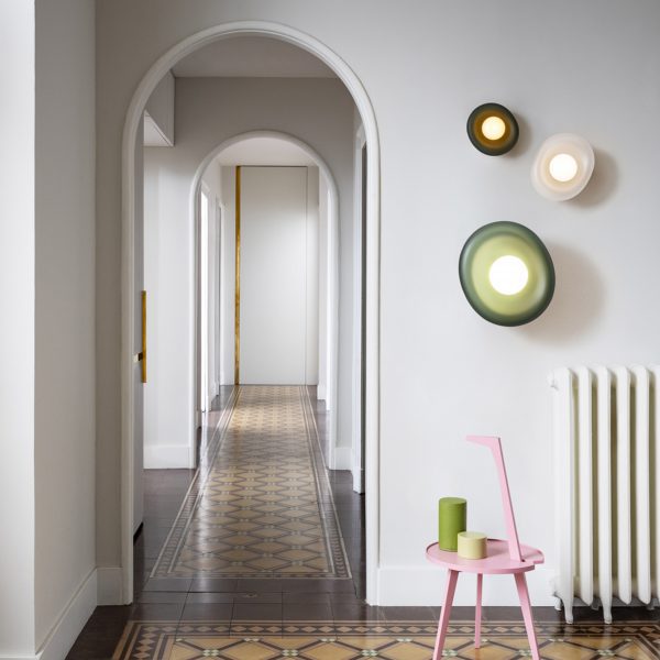 A photograph of a white wall with a hallway going through it, with a brown tiled floor. There are green, brown and white lights on the wall, above a small pink table.
