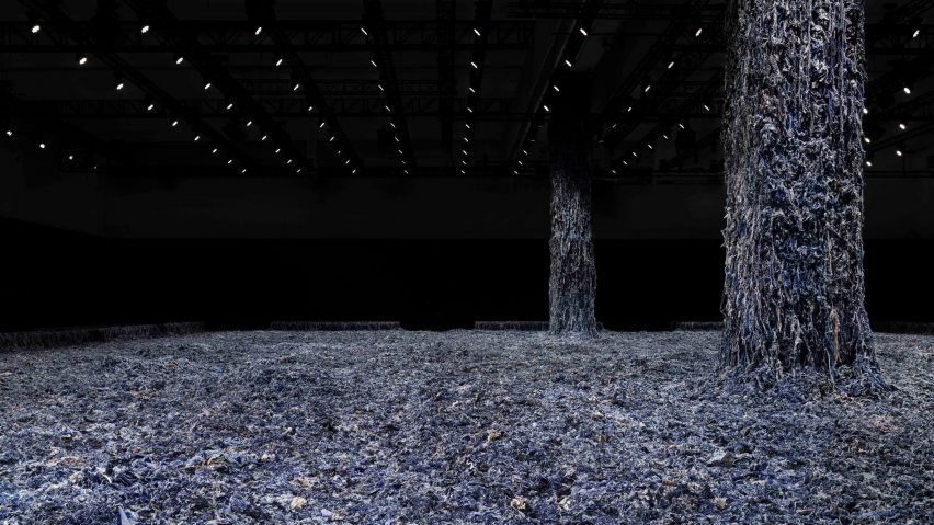A photograph of a large floorspace covered in blue denim scraps, with two columns emerging from it, also covered. The columns lead to a black ceiling with multiple lights in it.