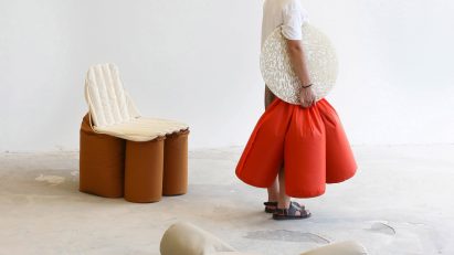 A photograph of a person, facing to the side, holding a seat featuring an orange cushion and a beige circular surface. There are two other seats surrounding them in tones of grey and brown.