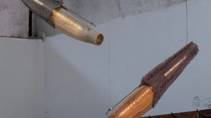 A photograph of two pendant lights, hanging diagonally, in tones of orange, beige and grey, against a white and grey concrete wall.