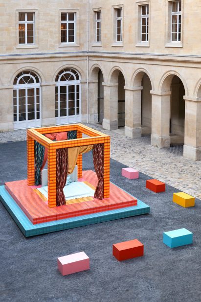 A photograph of an installation depicting a four-poster bed, in tones of orange, red, pink and blue, situated in a square in the Hôtel de la Marine.