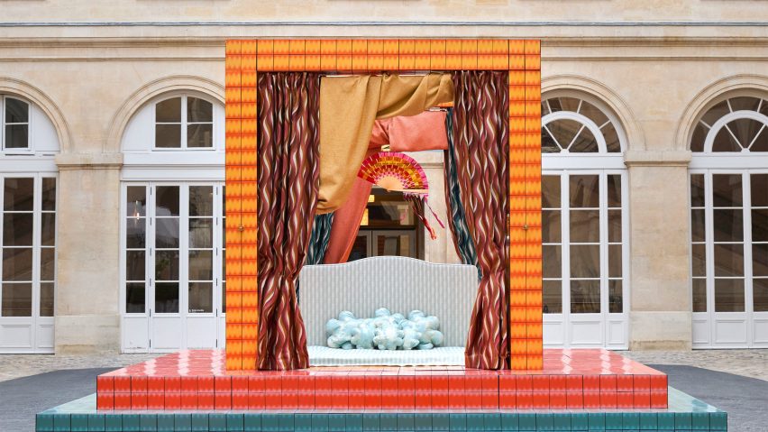 A photograph of an installation depicting a four poster bed in colours of blue, pink and orange, situated in a square of the Hôtel de la Marine.