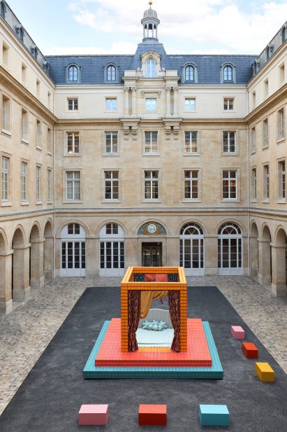A photograph of an installation depicting a four-poster bed, in tones of orange, red, pink and blue, situated in a square in the Hôtel de la Marine.