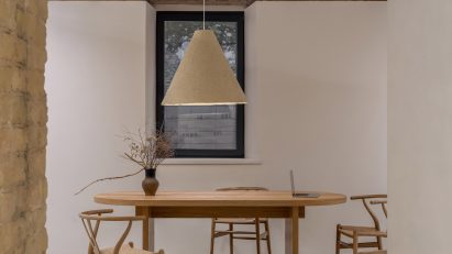 Photograph of a minimalist interior, showing a large wooden dining table with wishbone chairs and a large hanging pendant lamp.