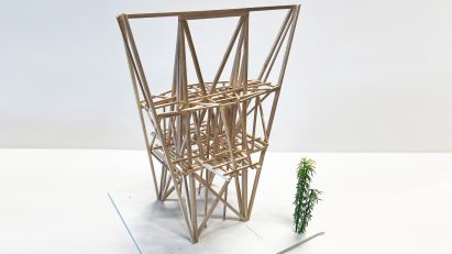 A photograph of an architectural model made from brown wooden sticks, in a geometric shape, against a white backdrop.