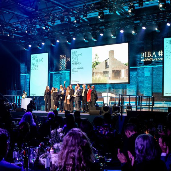 A photograph of an awards ceremony, showing people standing on a stage with screens behind them in tones of blue and white, and an audience of people sat in front of them.