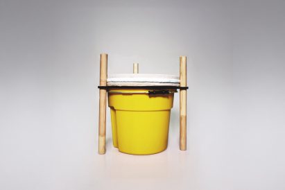 A photograph of a yellow bucket with brown wooden beams attached to it, against a grey backdrop.