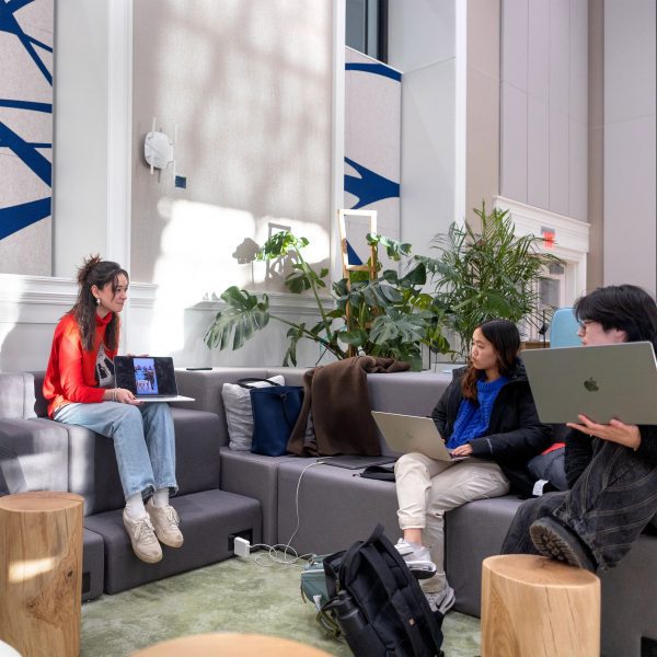 A photograph of three people with laptops sitting on benches, interacting with one another.