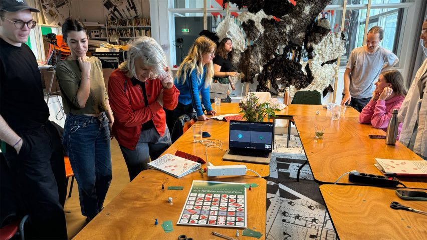 A photograph of people standing around a wooden table, interacting with one another.