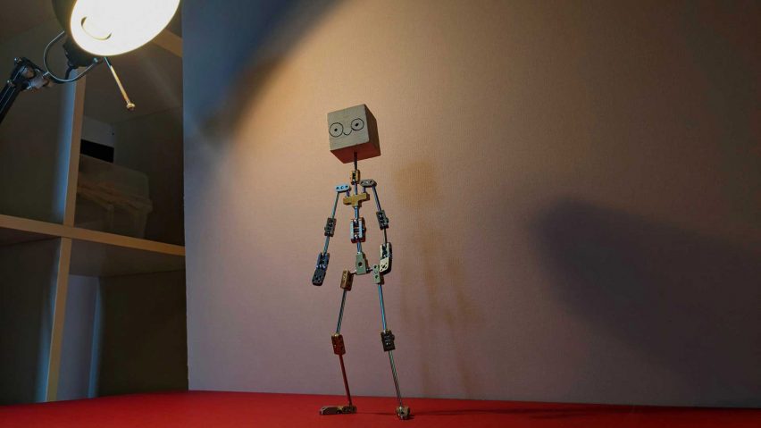 A 3D visualisation of a video game character, in tones of grey and beige with stick arms and legs, and a head in the shape of a cube. It is standing atop a red surface in front of a beige wall, with a light shining onto it.