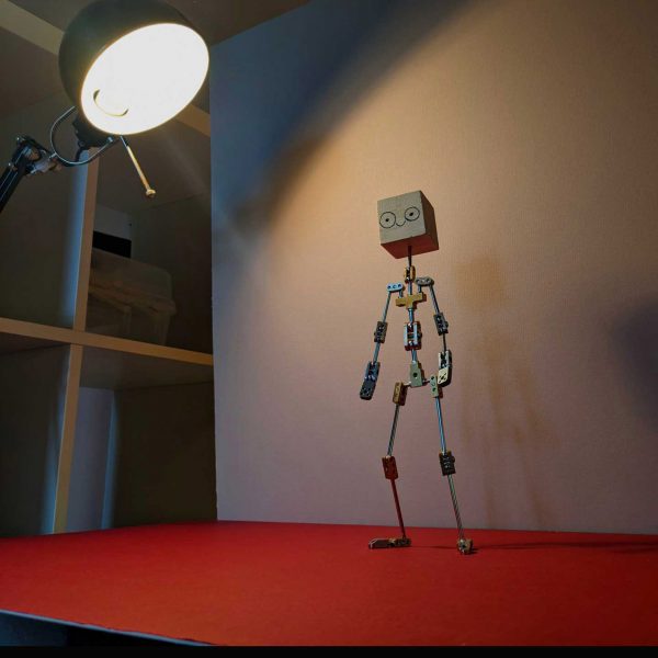 A 3D visualisation of a video game character, in tones of grey and beige with stick arms and legs, and a head in the shape of a cube. It is standing atop a red surface in front of a beige wall, with a light shining onto it.