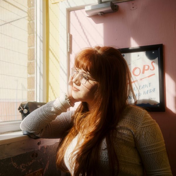A photograph of a person looking out of a window, with a pink wall behind them.