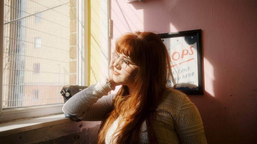 A photograph of a person looking out of a window, with a pink wall behind them.