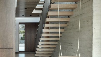 A photograph showing the underneath of a wooden floating staircase in tones of brown, with a white swing hanging from one of the stairs.