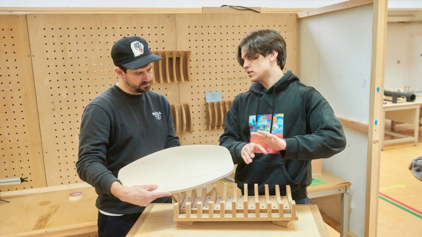 A photograph of two people interacting, in a wood workshop in tones of brown.