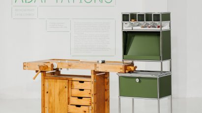 A photograph of a brown wooden workbench with a storage unit built into it. There is another storage unit beside it in tones of green, structured by silver beams. Behind them both is a white wall with green text on it.