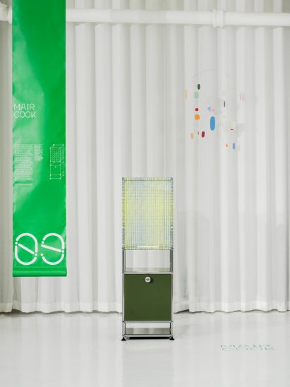 A photograph a rectangular lamp, structured with silver beams, with a yellow and blue textile covering and a dark green drawer at its base. There is a green rectangular banner to the left of it with white text on it.