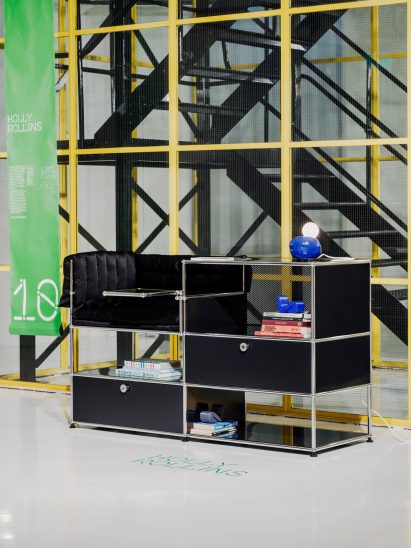 A photograph of a storage unit in tones of black and silver, against a glass wall with yellow beams.