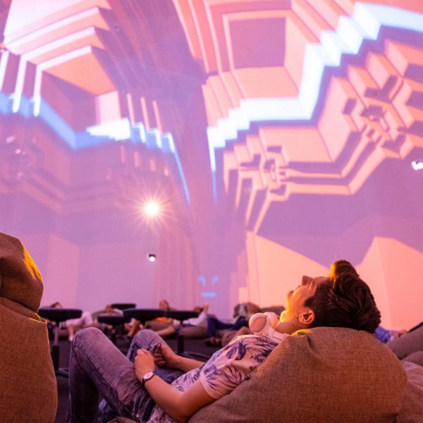 A photograph of a person lying down, looking up at a projection overhead in tones of pink and orange.