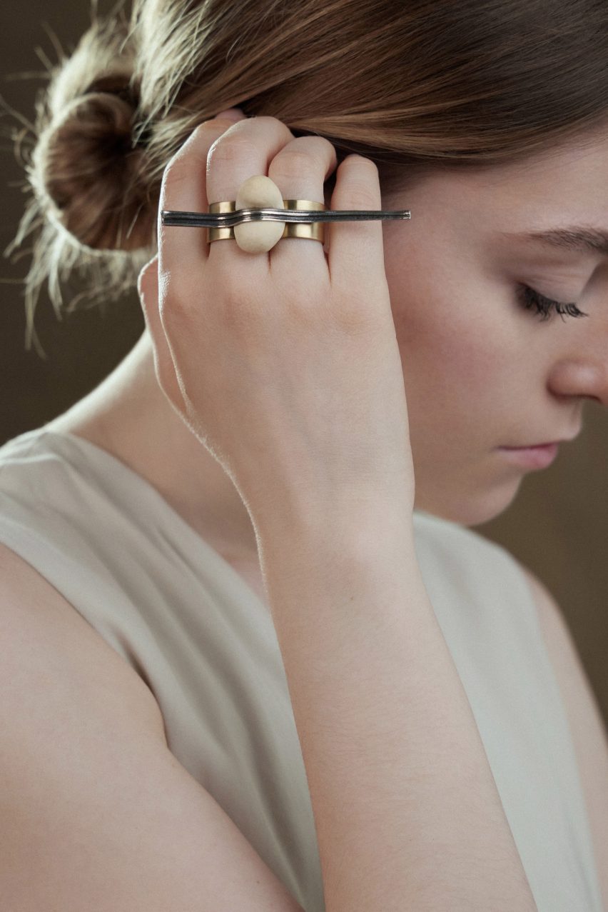 Close-up photo of a woman's head as she tucks hair behind her ear. She is wearing a chunky ring across two fingers that features a ball of baked dough flattened in the middle by a long, textured metal element that spans the width of the hand