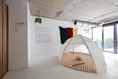 Photo of a white tent-like structure with a slatted timber base within a white exhibition space, surrounded by other objects