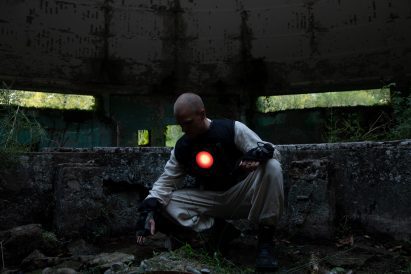 Photo of a man in loose beige clothing and a black vest with a glowing red disc over the heart crouching down to touch the earth in the middle of a ruined building overrun with plants