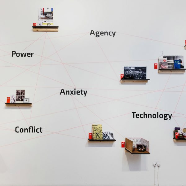 A photograph of a white wall with small shelves with various objects on them. There are multiple black words on the wall.