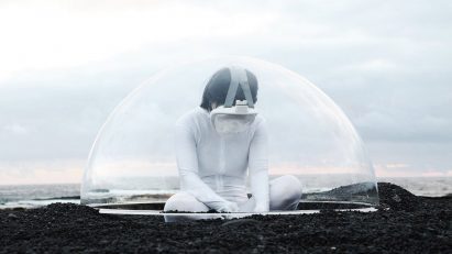 A photograph of a person in white clothing, with a VR headset on, sat on the ground in tones of brown. There is a translucent semi-sphere framing them against a pale blue backdrop.