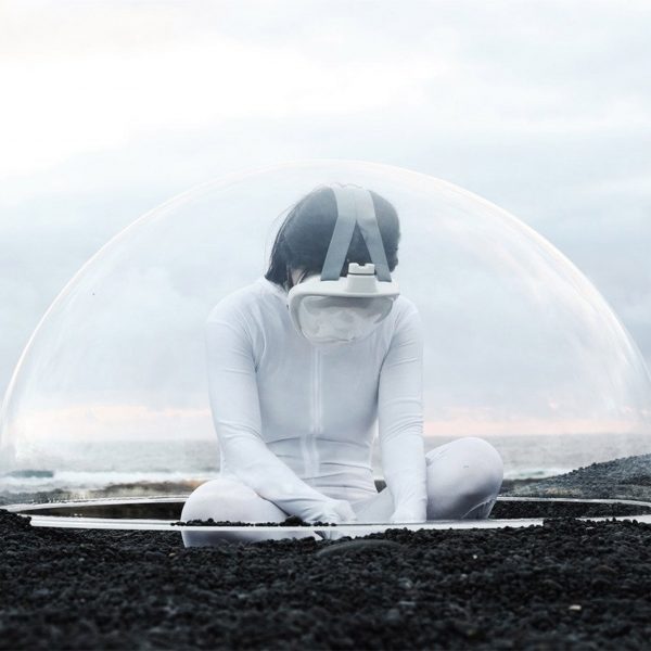 A photograph of a person in white clothing, with a VR headset on, sat on the ground in tones of brown. There is a translucent semi-sphere framing them against a pale blue backdrop.