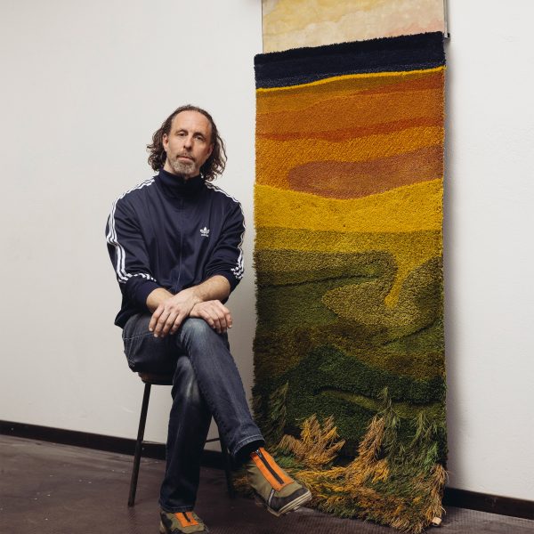 A photograph of a person sat next to an artwork which displays a gradient in tones of black, orange, yellow and green.