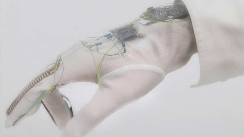 A close-up photograph of a person's hand, wearing a translucent white glove with blue wires and a black electrical device on it.