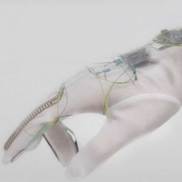 A close-up photograph of a person's hand, wearing a translucent white glove with blue wires and a black electrical device on it.