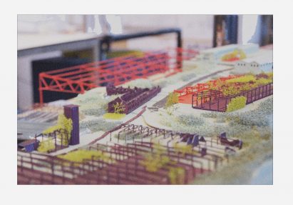A close up photograph of an architectural model, in tones of purple, red, green and white. There is a white border around the image.