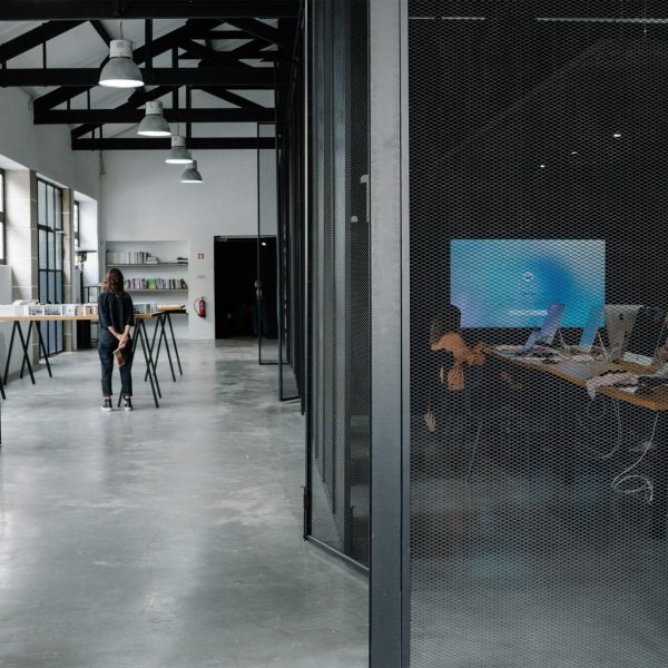 A photograph of a space in tones of grey. There is a workspace with computers to one side. There is also a table with a person standing next to it in the distance.