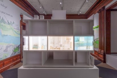 Close-up photo of the cross-section of an architectural model of an apartment building with on storey lit up and decorated with illustrations. The illustrations show furniture, books and other domestic items