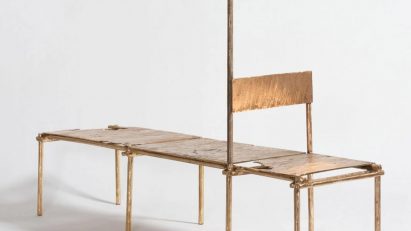 Studio photo of a daybed made from cast copper. At one end a backrest forms a chair, while a linear element extending vertically from the frame adds a candlestick from Oriki act II by Nifemi Marcus-Bello
