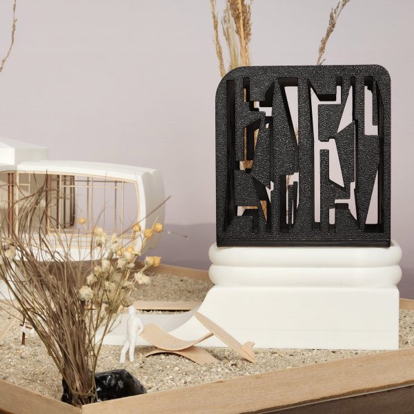 A photograph of an architectural model, in tones of white and black, atop a beige sanded surface.