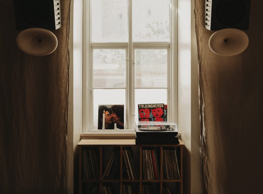 Photograph of a window framed by linen curtains with a vinyl cabinet and a turntable. On either side of the window hands two bespoke speakers.